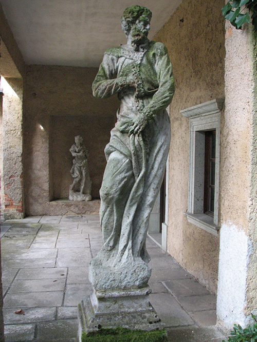 Escultura de una persona con barba y vestimenta antigua que sostiene un objeto alargado, ubicada en un pasillo con paredes de textura rugosa y otra escultura similar al fondo.

Descripción generada con IA
