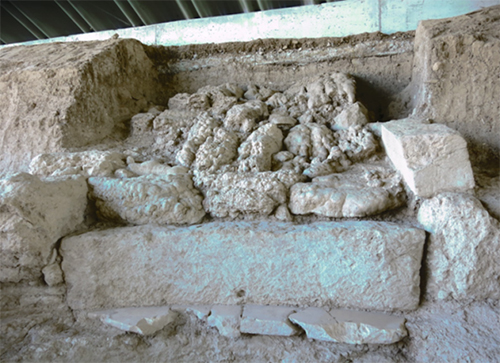 Restos arqueológicos de una estructura antigua de piedra y barro en un sitio excavado bajo un techo protector.

Descripción generada con IA