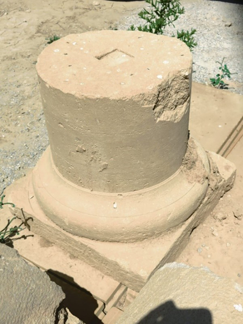 Restos arqueológicos de una columna de piedra cilíndrica con base cuadrada en un entorno arenoso al aire libre.

Descripción generada con IA
