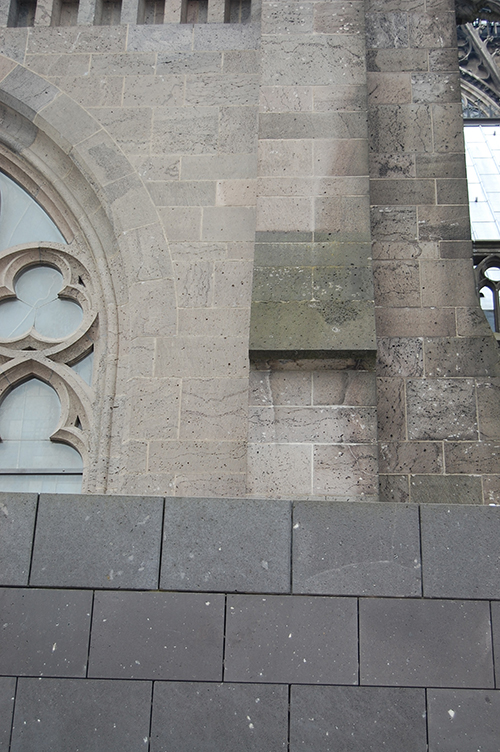 Parte de una fachada de piedra con una ventana gótica decorativa y un muro de bloques grises en primer plano.

Descripción generada con IA