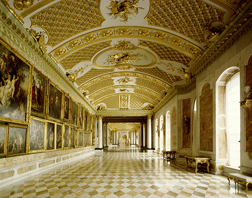 Pasillo largo y elegante con techo decorado en dorado, paredes adornadas con cuadros y ventanas altas que permiten la entrada de luz natural.

Descripción generada con IA