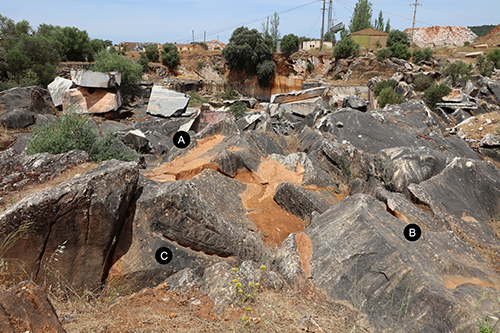 Un terreno rocoso con grandes piedras dispersas y vegetación baja en un área rural con casas y árboles al fondo bajo un cielo despejado.

Descripción generada con IA